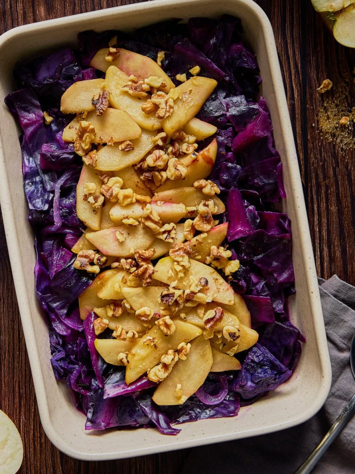 a serving dish on a wooden table filled with cabbage, apples, and walnuts