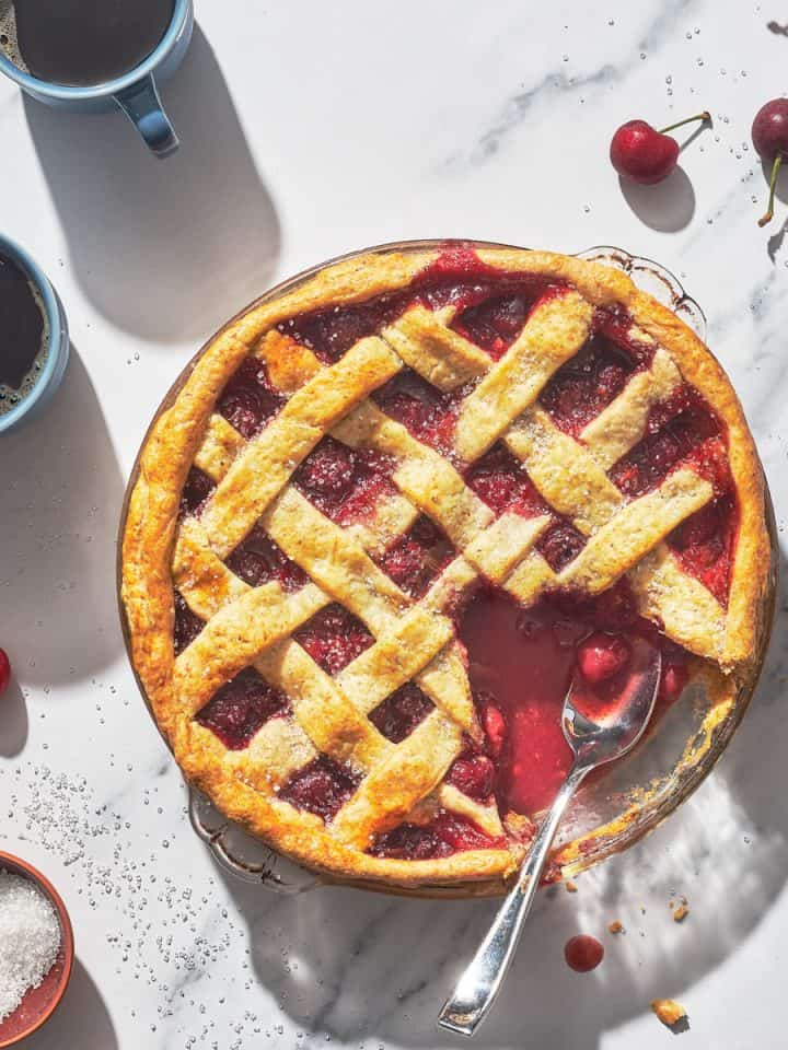a cherry rhubarb pie with a piece missing and coffee mugs