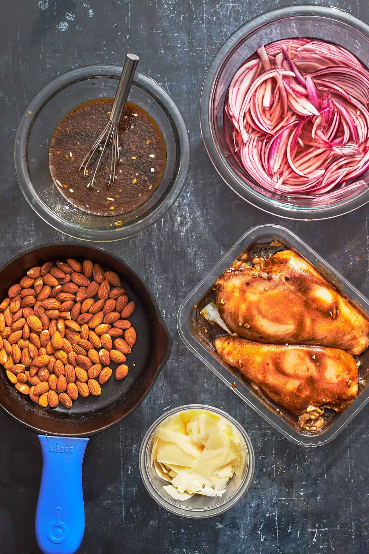 almonds toasting in small pan, bowl of onions, bowl of dressing, and chicken covered in marinade