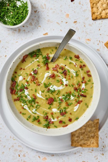 a bowl of asparagus soup with crackers on the side and bowls of chives and bacon