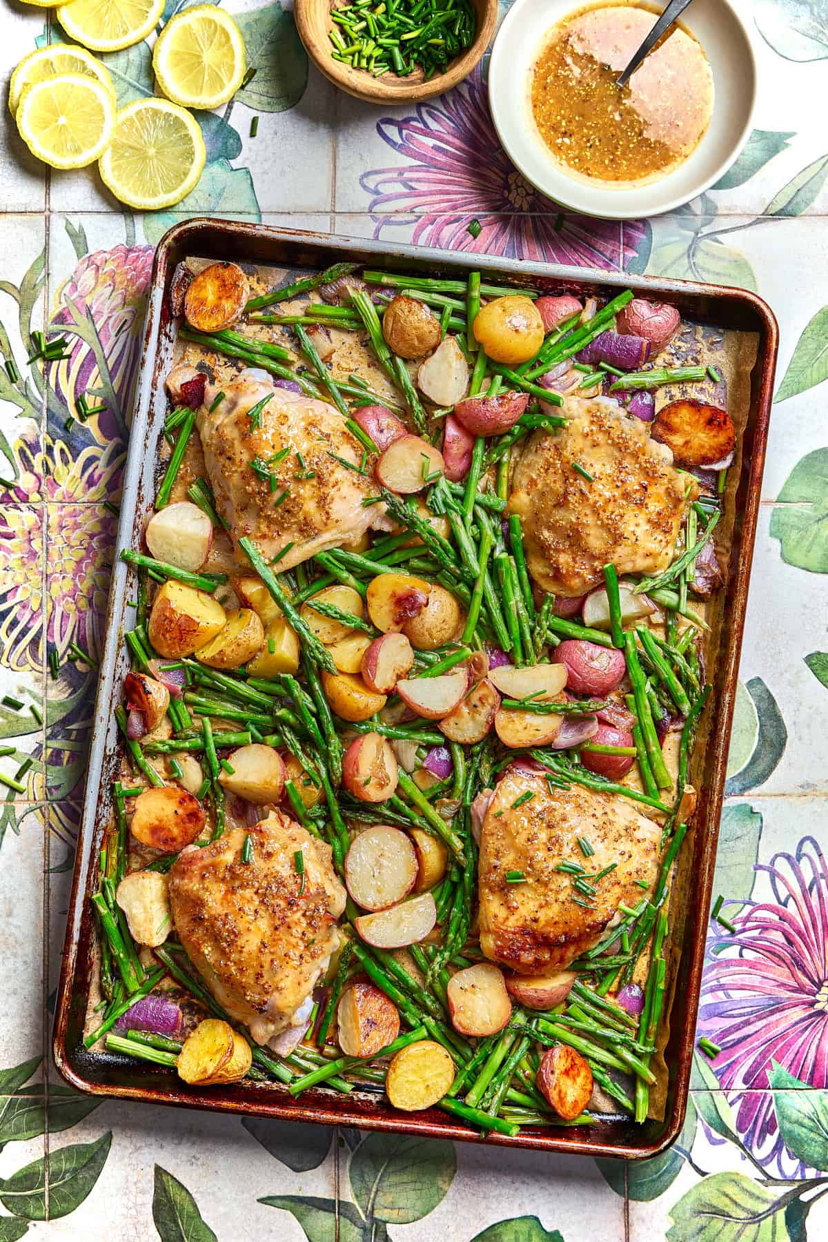 a sheet pan filled with chicken, potatoes, and asparagus with a dish of chives, slices of lemon, and a bowl of sauce on the side