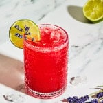 a glass filled with ice and a red mocktail garnished with lavender sprigs and a lime slice