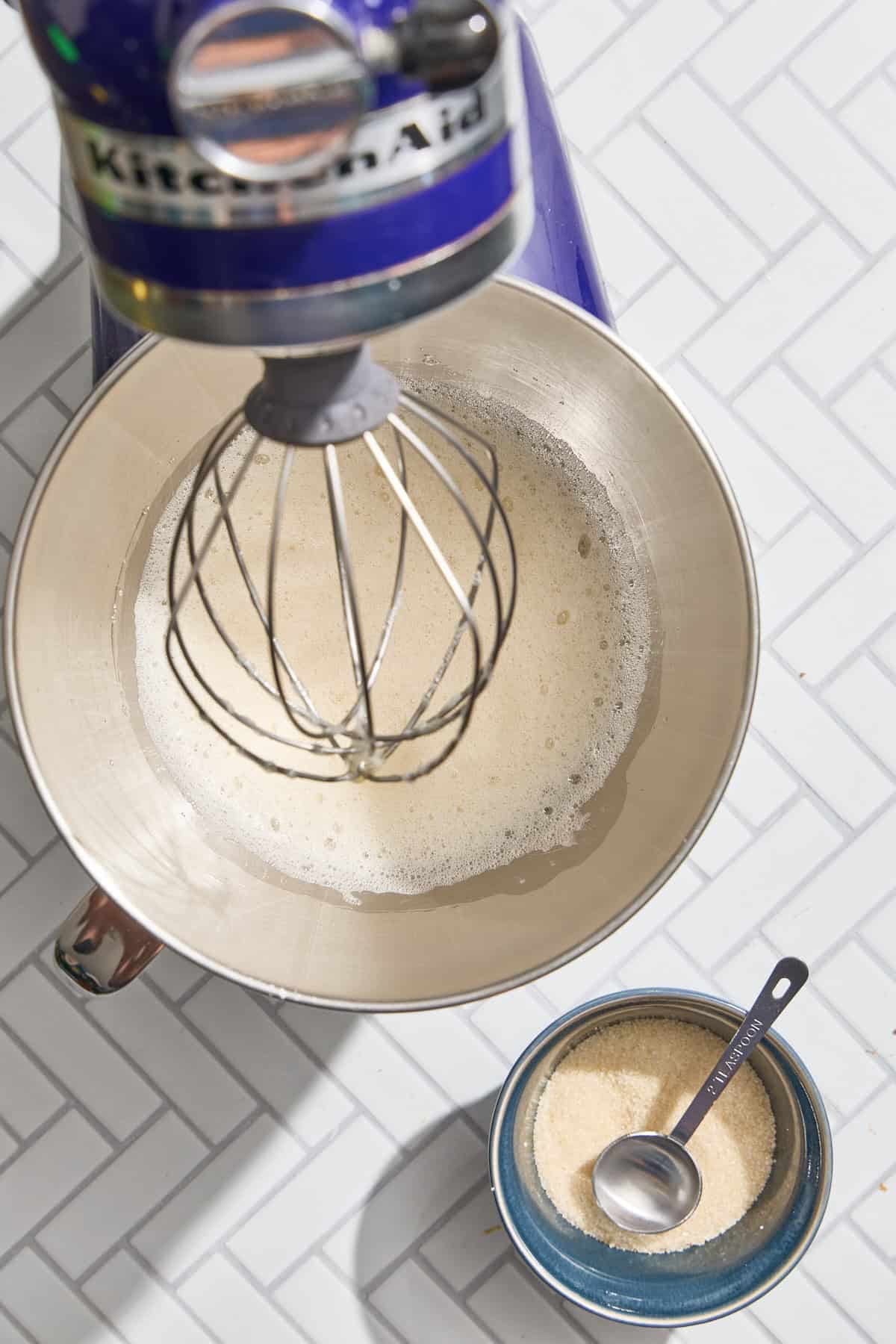 egg whites in a mixer with a small bowl of sugar