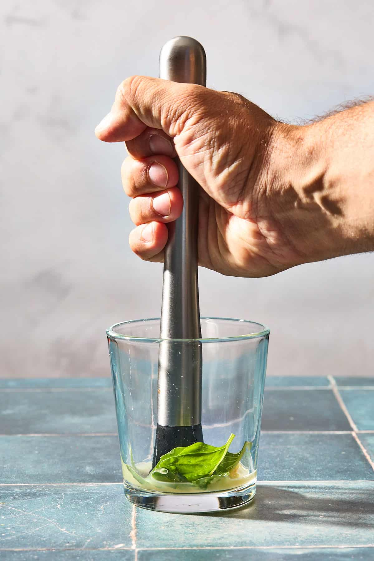 basil and liquid being muddled in a glass on a blue counter